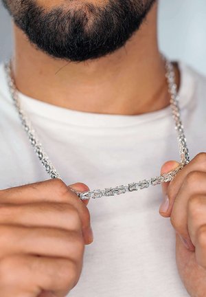 Silberne Kettenhalskette mit ineinander greifenden Gliedern, die zwischen den Fingern gehalten wird. Getragen über einem schlichten weißen Hemd, mit einer glatten, polierten Oberfläche.