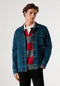 Giubbotto di jeans blu scuro con una vestibilità comoda, caratterizzato da una tasca frontale, bottoni metallici e cuciture a vista, indossato sopra un maglione a righe.