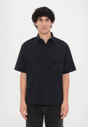 Camicia corta da uomo di colore navy realizzata in tessuto testurizzato. Presenta una chiusura con bottoni sul davanti, una tasca sul petto e un accento in pelle marrone.