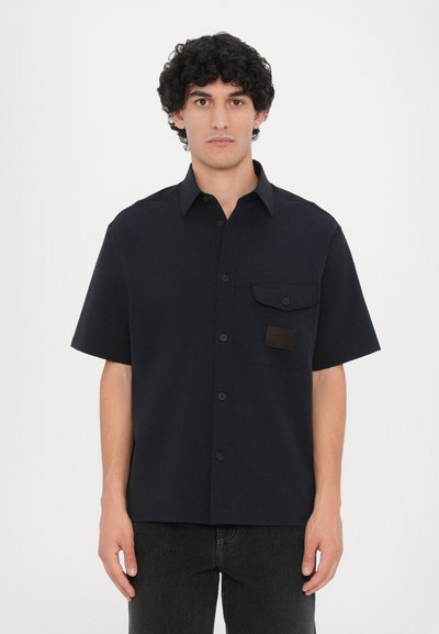 Marineblauw kortmouwshirt van textuurstof. Voorzien van een knoopsluiting aan de voorkant, een borstzak en een bruin leren patchaccent.