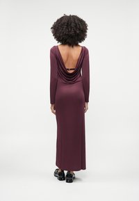 Femme aux cheveux bouclés portant une robe bordeaux à manches longues et longue jusqu'au sol, avec un dos drapé bas et des chaussures en cuir verni noires, debout sur un fond blanc.