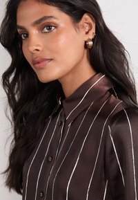 Braun gestreiftes Hemd mit Kragen, glänzendem Stoff und schwarzen Knöpfen. Modell mit langen Haaren trägt goldene Creolen.