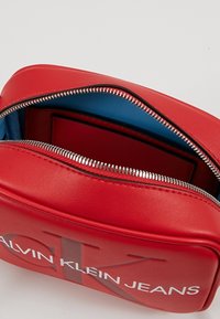 Červená taška Calvin Klein Jeans se stříbrným zipem, částečně otevřená, ukazuje modrou vnitřní podšívku.