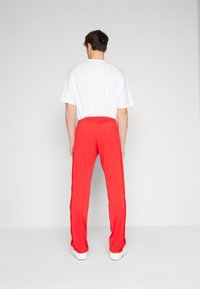 Muž v jasně červených sportovních kalhotách a jednoduchém bílém tričku stojí s pohlavím k publiku na jednobarevném světlém pozadí.