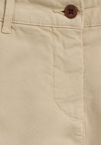 I pantaloni in cotone beige presentano una texture liscia, una lieve elasticità diagonale, una tasca frontale e un bottone rotondo marrone in vita.