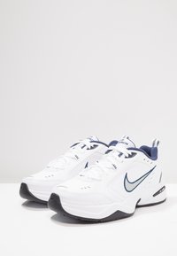 Zapatillas deportivas blancas con acentos en azul marino, con una parte superior sintética, cuello acolchado, cordones y un diseño de suela negra contrastante.