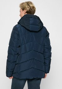 Eine Person, die eine dunkelblaue gesteppte Jacke mit Kapuze trägt, von hinten gesehen, vor einem schlichten hellen Hintergrund stehend.