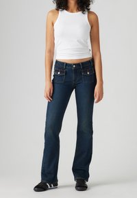 Jean en denim bleu foncé avec une taille cintrée et des jambes évasées, doté de deux poches avant avec fermetures par boutons. Associé à un débardeur blanc.