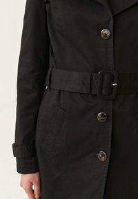 Czarny płaszcz o długości do kolan ma pasek w talii, duże guziki i boczne kieszenie. Tkanina ma gładką, matową teksturę.