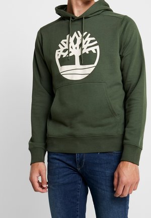 Mężczyzna w zielonej bluzie z kapturem z dużym białym logo drzewa na klatce piersiowej i niebieskich dżinsach, ukazany od ramion do ud.