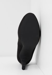 Chaussure noire à talons hauts avec une surface texturée, un bout pointu et une semelle en plastique lisse. Le talon est fin et mesure environ dix centimètres de hauteur.