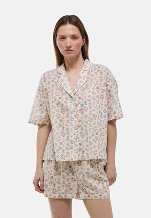 Femme portant un ensemble pyjama à manches courtes de couleur claire avec une chemise boutonnée et un short assorti, debout devant un fond uni.