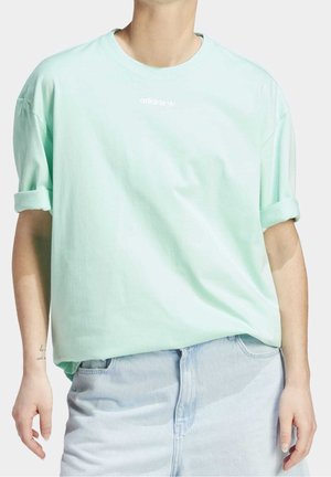 Camiseta de algodón de color verde menta de gran tamaño con mangas enrolladas y logo blanco de "adidas" en el pecho, combinada con una falda de mezclilla azul clara.
