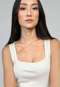 Débardeur à côtes sans manches en blanc cassé avec un décolleté carré. Présente une discrète marque et une coupe ajustée qui met en valeur la silhouette.