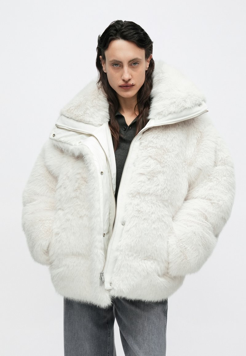 Veste oversize en fausse fourrure blanche avec un col montant, une fermeture éclair et de grandes poches, présentant une finition douce et texturée ainsi qu'une doublure lisse.