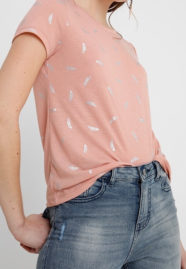 Femme portant un t-shirt rose à manches courtes avec des motifs de feuilles argentées rentrée dans un jean bleu délavé.