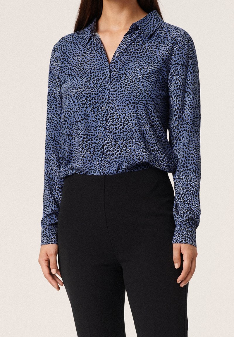 Femme portant une chemise bleue à motifs à manches longues, boutonnée et rentrée dans un pantalon noir taille haute, debout devant un fond uni.