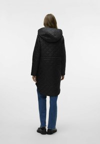 Zwarte gewatteerde lange jas met een kap, met een middellange ontwerp, zijventilaties en een gladde textuur. Draagt over blauwe straight-leg jeans.