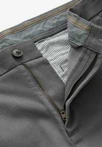 Pantalon gris texturé avec fermeture éclair en laiton non fermée, ceinture boutonnée, et doublure intérieure rayée visible à l'ouverture de la taille.