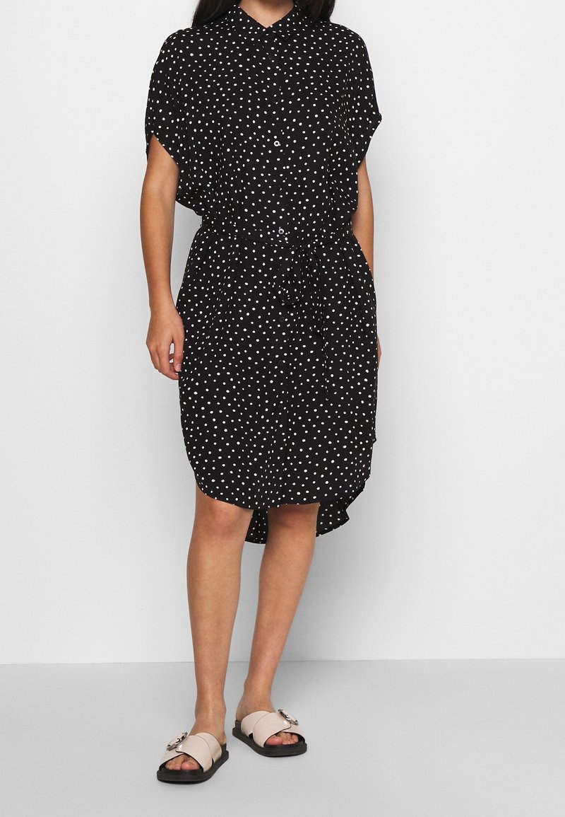 Femme portant une robe noire longueur genou à pois blancs, boutonnée sur le devant, manches courtes, ceinture à nouer à la taille et sandales beiges à enfiler.