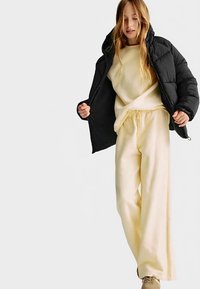 Czarna puchowa kurtka z kapturem, beżowa bluza oraz jasnobeżowe spodnie dresowe. Gładki materiał, luźny krój i szczegóły z elastycznym pasem.