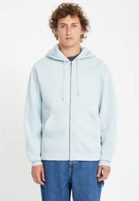 Hoodie zippé bleu clair en tissu doux, avec une poche kangourou à l'avant et une capuche avec cordon de serrage, associé à un jean bleu ample.
