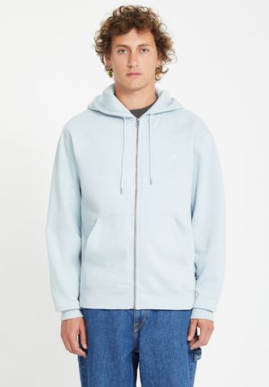 Sudadera con cremallera de color azul claro en tejido suave, con un bolsillo tipo canguro en la parte frontal y capucha con cordón, combinada con jeans azules holgados.