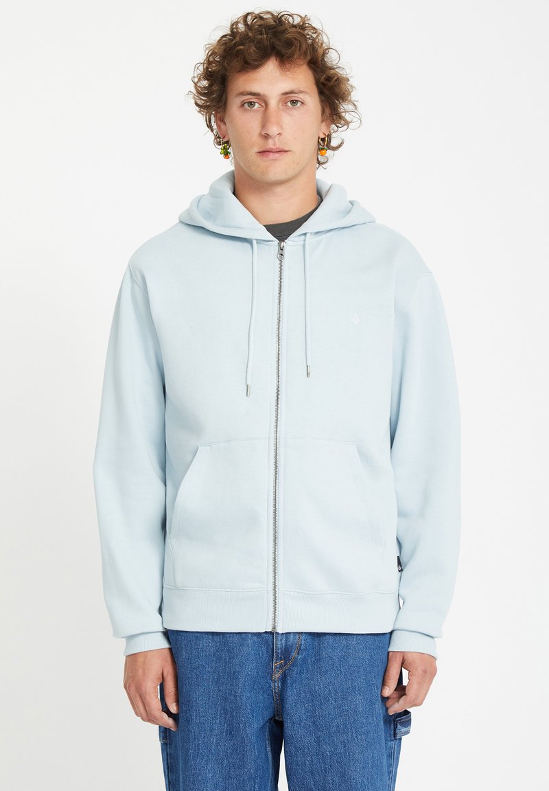 Hoodie zippé bleu clair en tissu doux, avec une poche kangourou à l'avant et une capuche avec cordon de serrage, associé à un jean bleu ample.