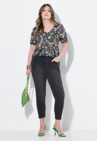 Top negro con patrón floral y escote en V, combinado con jeans negros ajustados que presentan un costura lateral decorativa. Lleva tacones de aguja verdes y un clutch.
