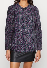 Blusa floreale con maniche lunghe, sfondo scuro, fiori rosa e verdi, chiusura con bottoni, tessuto testurizzato, abbinata a una gonna di pelle nera.