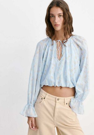 Mujer con cabello castaño ondulado que lleva una blusa de manga larga azul claro con flores y puños con volantes, y pantalones beige de cintura alta, de pie frente a un fondo simple.
