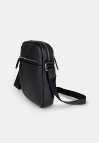 Černá kožená crossbody taška s texturovaným povrchem, obsahující dvojité zipy, plochou přední kapsu a nastavitelné látkové ramínko.