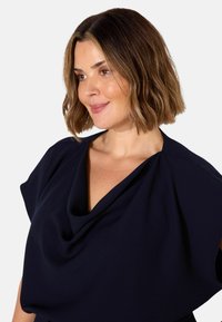 Top color blu navy con scollatura drappeggiata, maniche corte e texture di tessuto morbida. Presenta una vestibilità comoda e una finitura liscia.