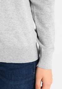 Šedý svetr z měkké pleteniny s žebrovanými manžetami a lemem, zobrazený vedle tmavých džínů z denimu. Viditelné detailní šití.