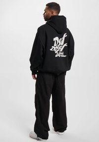 Svart hettegenser med hvitt grafisk tekst på baksiden. Løs passform med hette, kombinert med svarte baggy bukser og hvite sneakers.