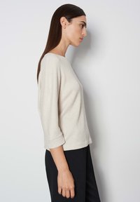 Hellbeige strukturierte Bluse mit Bootsausschnitt, Dreiviertelärmeln und umgeschlagenen Manschetten, kombiniert mit schwarzen Hosen.