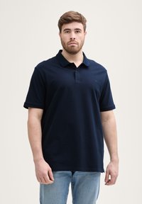 Tamsiai mėlynos spalvos polo marškiniai, pagaminti iš medvilnės, su apykakle, trumpomis rankovėmis ir subtiliu logotipu ant krūtinės. Suderinti su šviesiai mėlynomis džinsais.