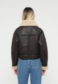 Mujer de espaldas con chaqueta de piel de oveja marrón oscuro con cuello y puños beige, combinada con vaqueros de cintura alta azules, contra un fondo blanco.