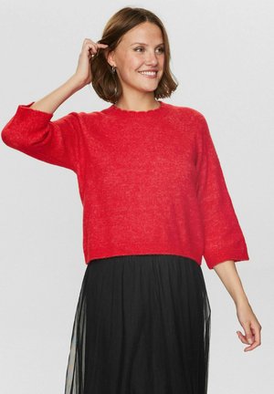 Suéter rojo de punto con cuello redondo, mangas tres cuartos y dobladillo con puntilla. Combinado con una falda negra, fluida, para crear contraste.