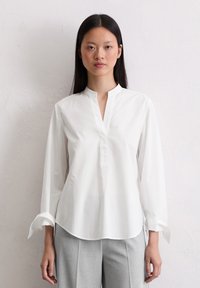 Blusa bianca in cotone con colletto alla cinese, maniche lunghe e patta frontale parziale. Presenta un orlo curvo e polsini arrotolati.