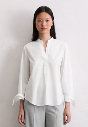 Blouse en coton blanc avec un col mandarin, des manches longues et une patte de boutonnage partielle sur le devant. Présente un ourlet arrondi et des poignets retroussés.