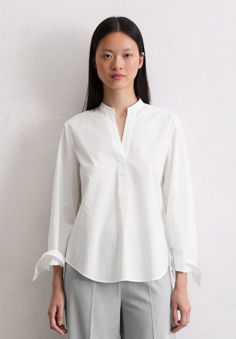 Blusa bianca in cotone con colletto alla cinese, maniche lunghe e patta frontale parziale. Presenta un orlo curvo e polsini arrotolati.