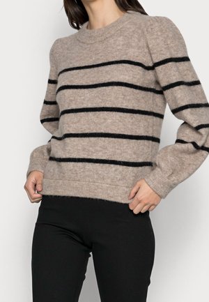 Vrouw draagt een beige zachte trui met zwarte horizontale strepen en een zwarte broek, handen houden de zoom van de trui vast.