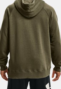 Homme portant un hoodie vert olive avec des poignets et un ourlet côtelés, présenté de dos, associé à un pantalon noir avec un motif blanc.
