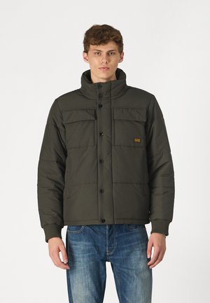 Padded olivgrüne Jacke mit hohem Kragen, vorderen Druckknöpfen und zwei Brusttaschen. Über blauen Jeans getragen.