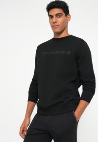 Črna sweatshirt s krojnim ovratnikom in rebrastimi manšetami, ki ima reliefni logotip "KARL LAGERFELD" čez prsa. Gladka tekstura, sproščen kroj.