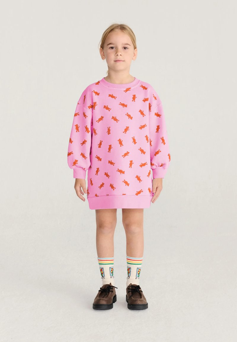 Enfant debout portant une robe rose avec des imprimés de chiens orange, des chaussettes colorées à motifs et des chaussures marron sur un fond clair uni.