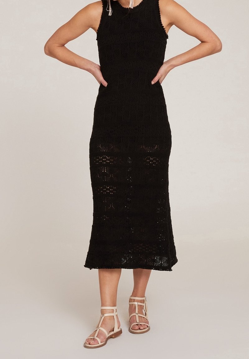 Femme debout, mains sur les hanches, portant une robe midi noire en crochet sans manches et des sandales plates beige à lanières sur un fond neutre.