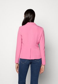 Kobieta z długimi ciemnymi włosami, ubrana w dopasowaną różową marynarkę i niebieskie dżinsy, stojąca przodem do białego tła.