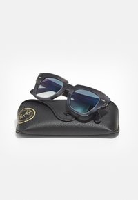 Ochelari de soare Ray-Ban negri, cu o ramă pătrată, lentile cu degrade albastru și accente hardware subtile, așezați pe un toc texturat negru.